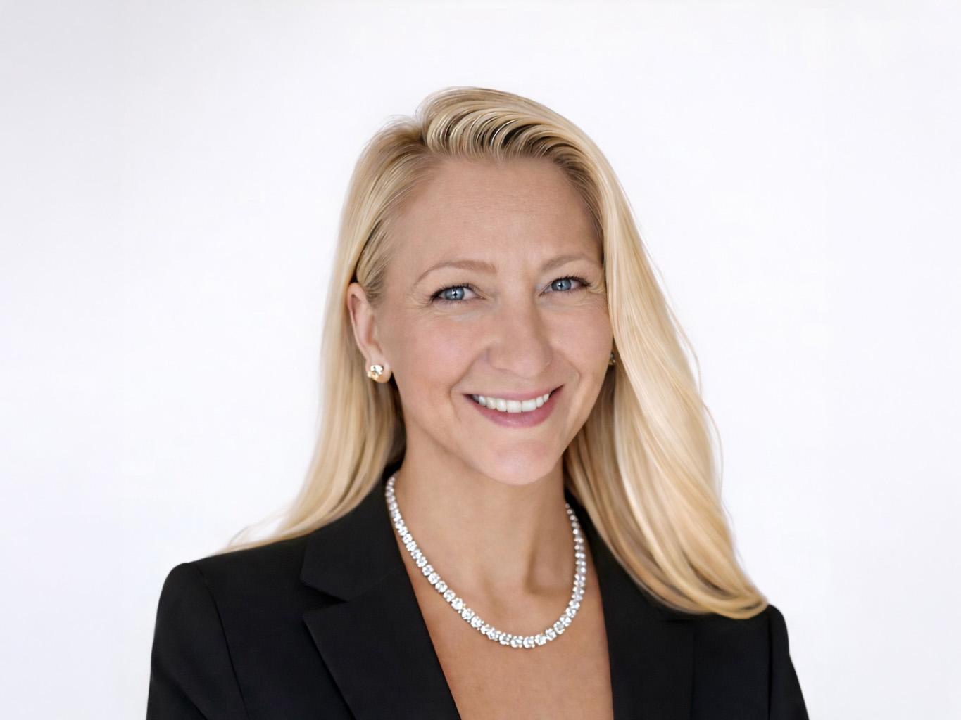 A blond woman with blue eyes wearing a diamond necklace and a black blazer smiles for a portrait. 
