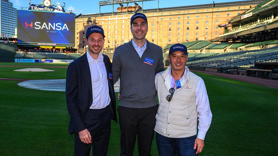 Three men who co-founded and lead Huxley Medical stand together on the field of a baseball stadium, smiling at the camera. They are wearing SANSA-branded hats, with a large video board behind them displaying the SANSA logo and the stadium seating in the background.