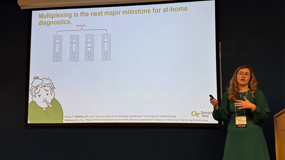 Presenter giving a talk on multiplexing for at‑home diagnostics, standing beside a slide showing test strip illustrations and scientific text.